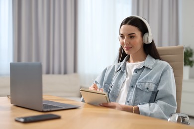 Photo of Young woman in headphones watching webinar at table in room