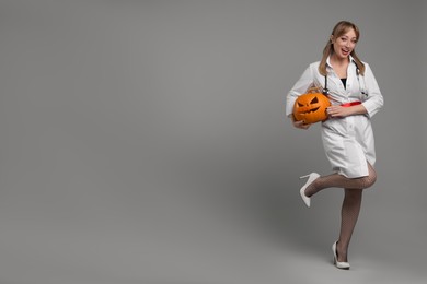 Photo of Happy woman in scary nurse costume with carved pumpkin on light grey background, space for text. Halloween celebration