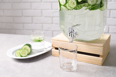 Photo of Jar dispenser of fresh cucumber water and glasses on table against brick wall. Space for text