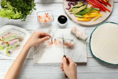 Woman cutting rice paper roll at white wooden table, top view