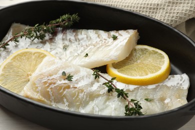 Fresh raw cod fillets with thyme and lemon in baking dish on table, closeup