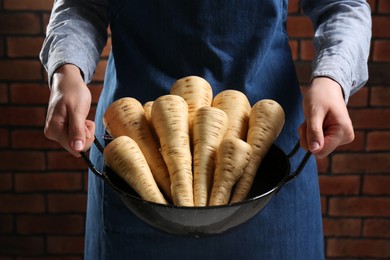 Photo of Woman holding bowl with fresh ripe parsnips near red brick wall, closeup