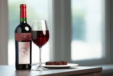 Photo of Bottle and glass of red wine with chocolate candies on wooden table indoors
