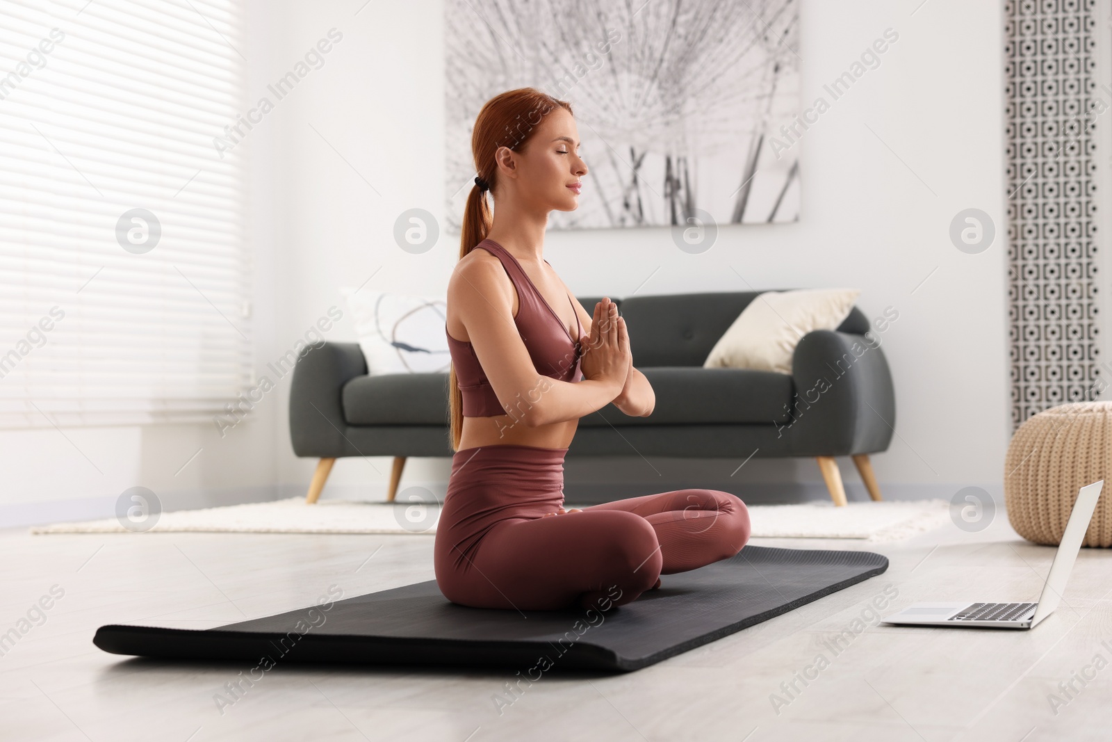 Photo of Beautiful young woman practicing Padmasana with laptop on yoga mat at home. Lotus pose