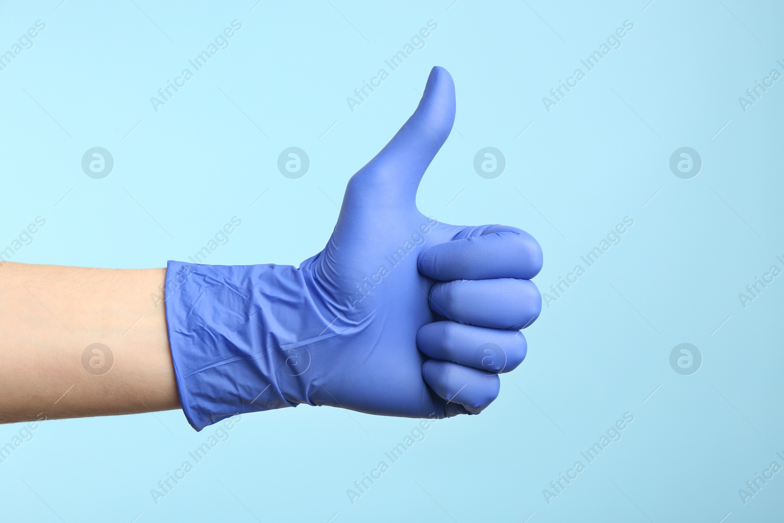 Photo of Person in medical gloves showing thumb up on light blue background, closeup of hand