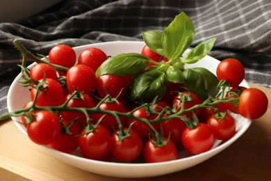 Photo of Plate of ripe whole cherry tomatoes with water drops and basil leaves on wooden table, closeup