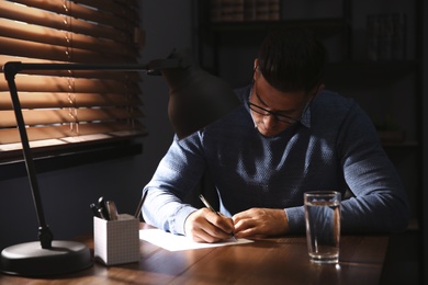 Photo of Man writing letter at wooden table in dark room