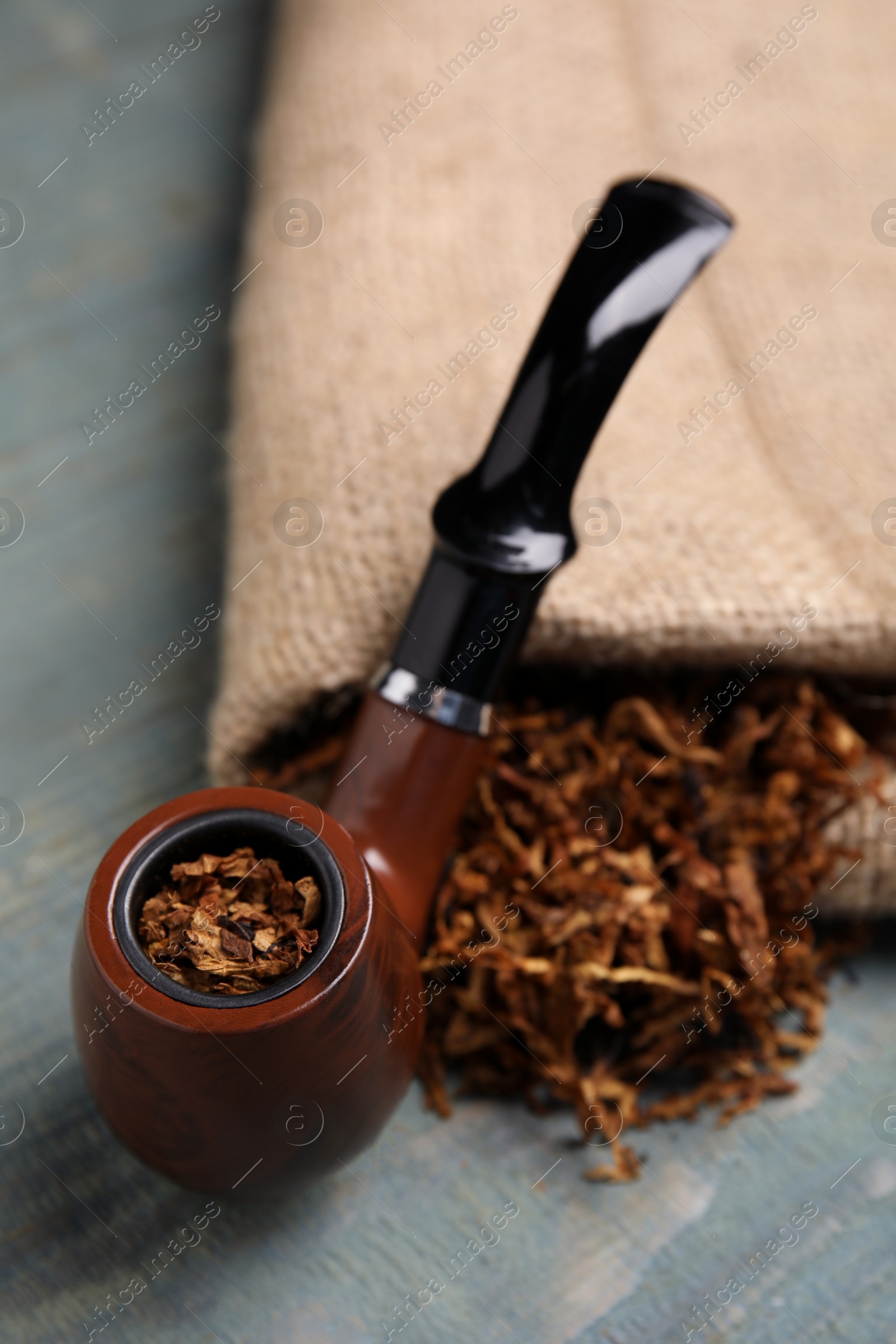 Photo of Smoking pipe, dry tobacco and sackcloth on blue wooden table, closeup