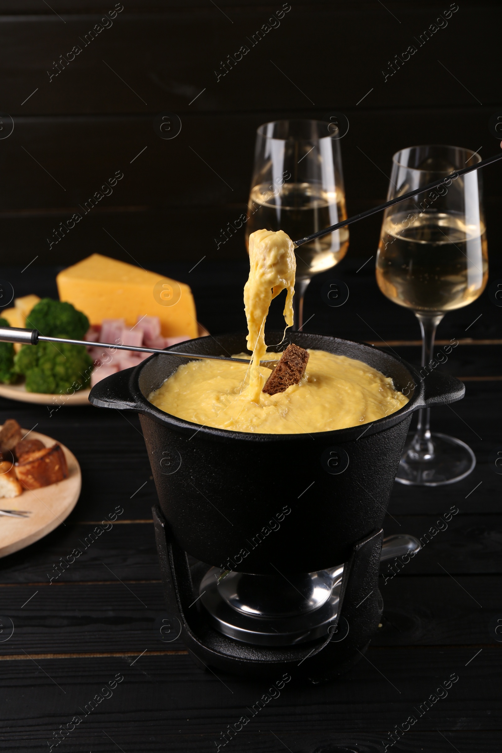 Photo of Dipping pieces of ham and bread into fondue pot with melted cheese on black wooden table