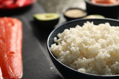 Photo of Cooked rice in bowl and fresh salmon for sushi on dark table, closeup