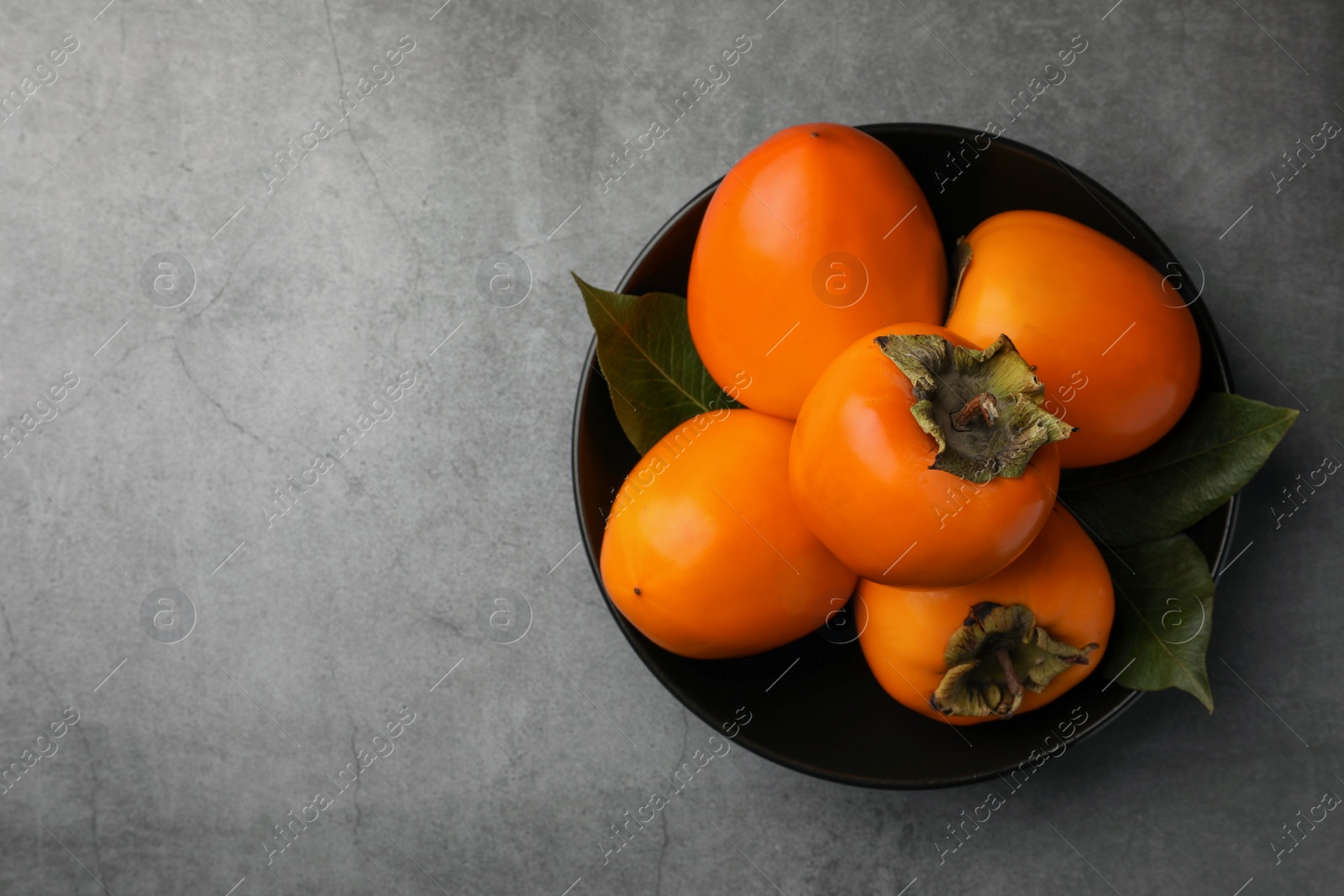 Photo of Delicious ripe persimmons in bowl on light gray textured table, top view. Space for text