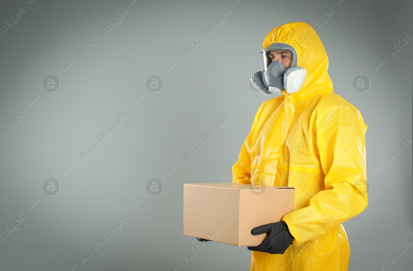 Photo of Man wearing chemical protective suit with cardboard box on light grey background, space for text. Prevention of virus spread