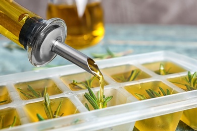 Photo of Pouring olive oil into ice cube tray with rosemary on table, closeup