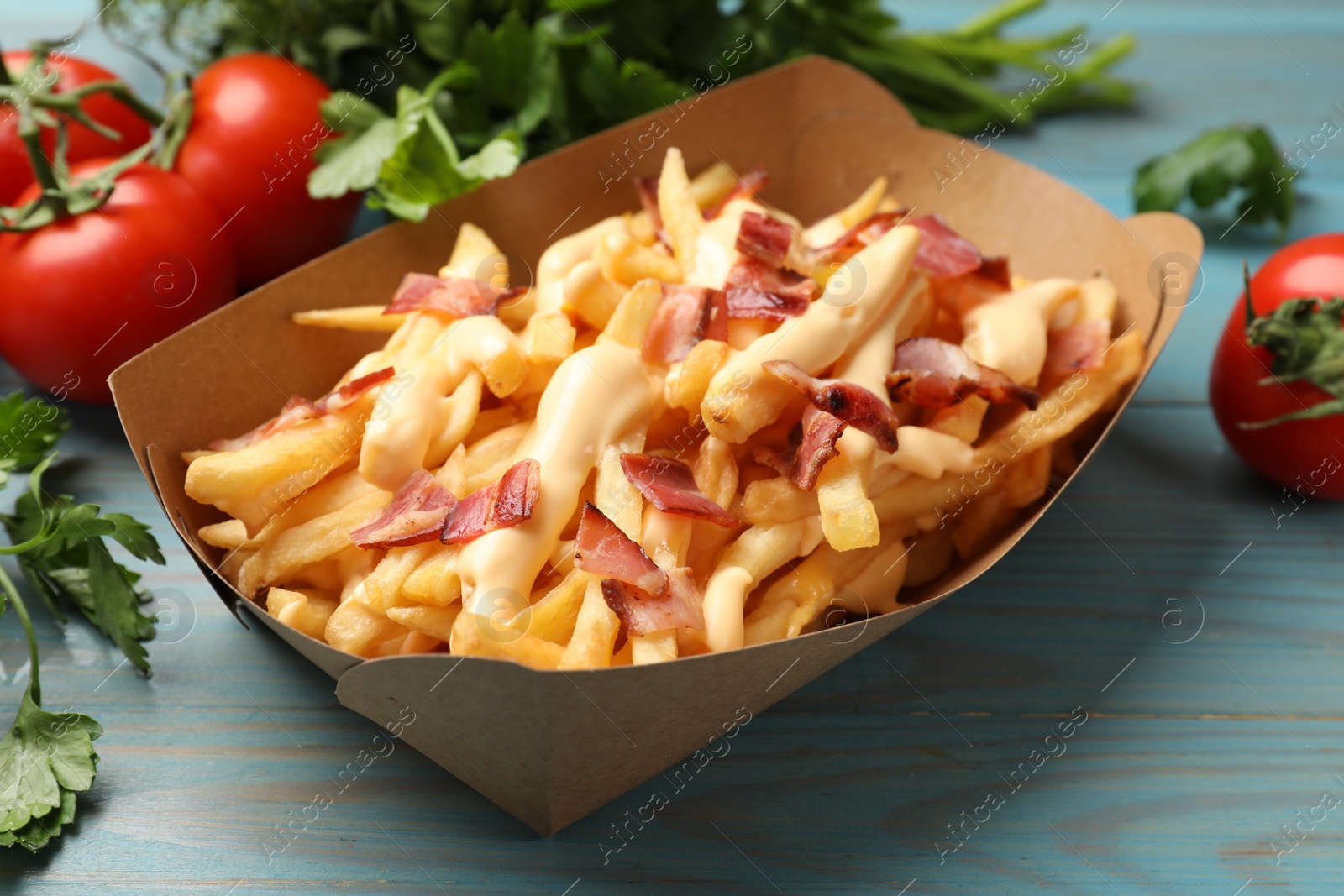 Photo of Tasty potato fries, cheese sauce, bacon and products in paper container on light blue wooden table, closeup