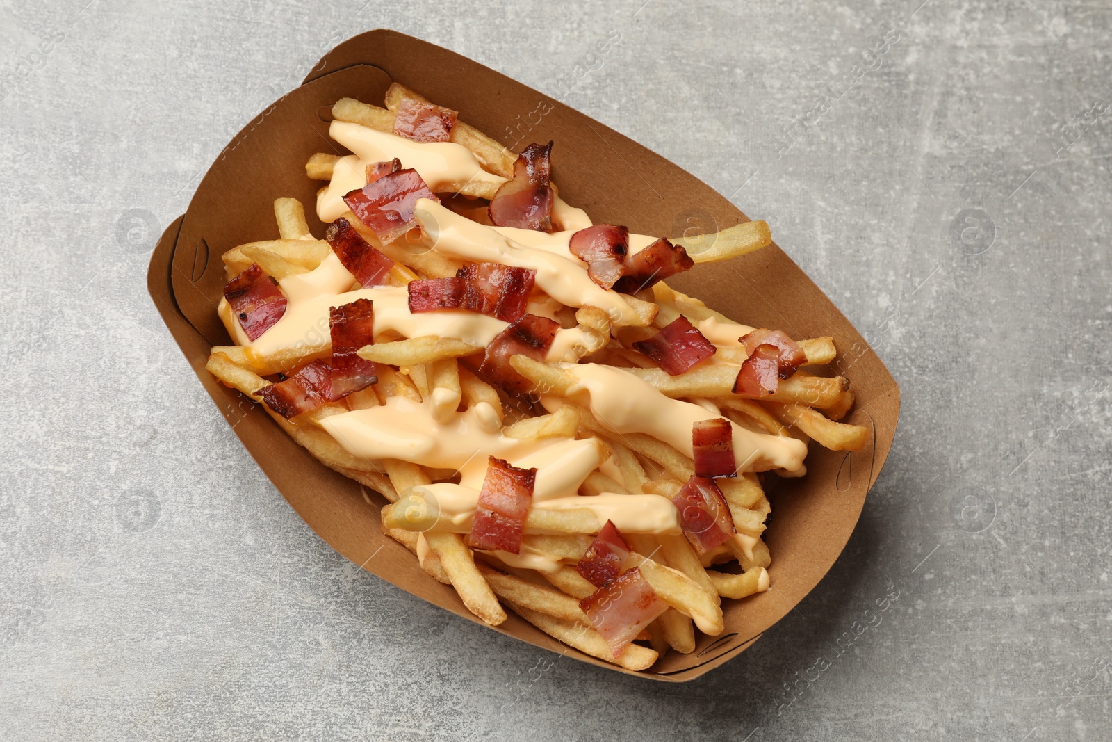 Photo of Tasty potato fries, cheese sauce and bacon in paper container on grey table, top view