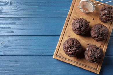Photo of Delicious chocolate muffins and sieve with cocoa powder on blue wooden table, top view. Space for text