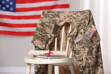 Chair with soldier uniform, notebooks and diploma near flag of United States indoors. Military education