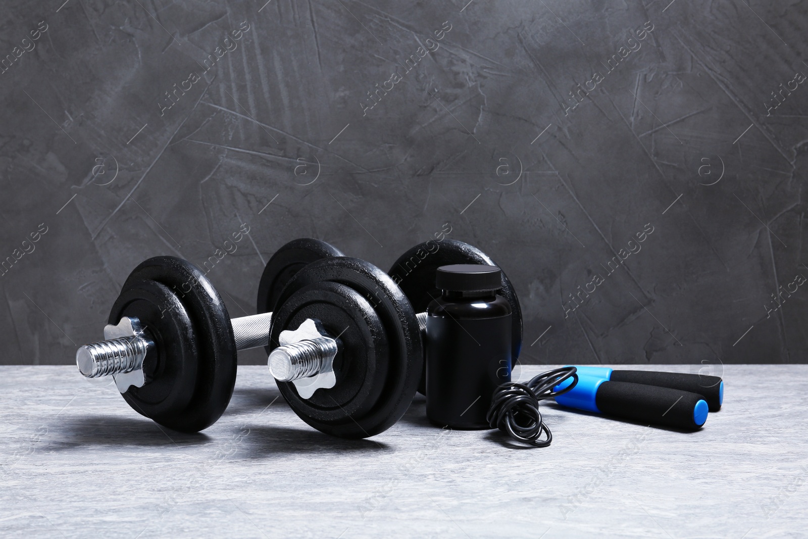 Photo of Pair of adjustable dumbbells, jump rope and jar with protein on floor near gray wall. Space for text