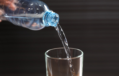 Photo of Pouring water from bottle into glass against dark background, closeup