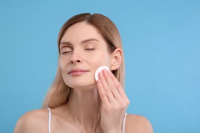 Beautiful woman removing makeup with cotton pad on light blue background, space for text