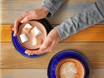 Woman holding cup of aromatic cacao with   marshmallows at wooden table, top view