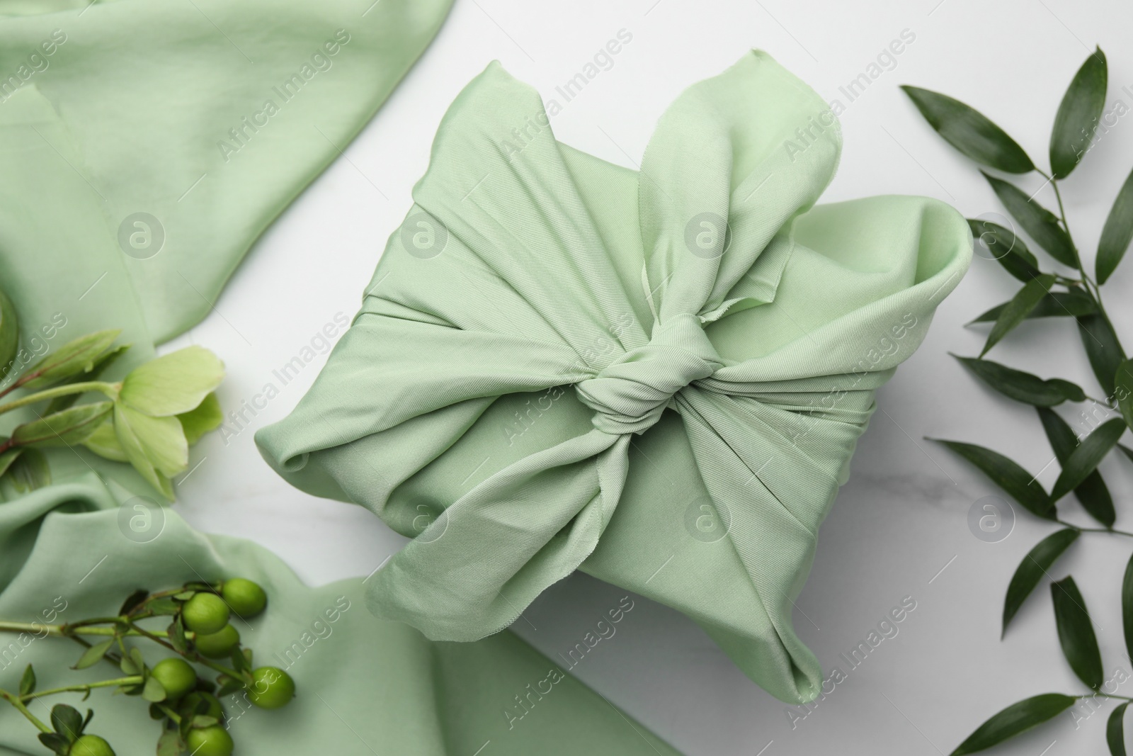 Photo of Furoshiki technique. Gift packed in green fabric and plants for decor on white table, flat lay