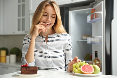 Photo of Woman choosing between cake and healthy fruits at table in kitchen