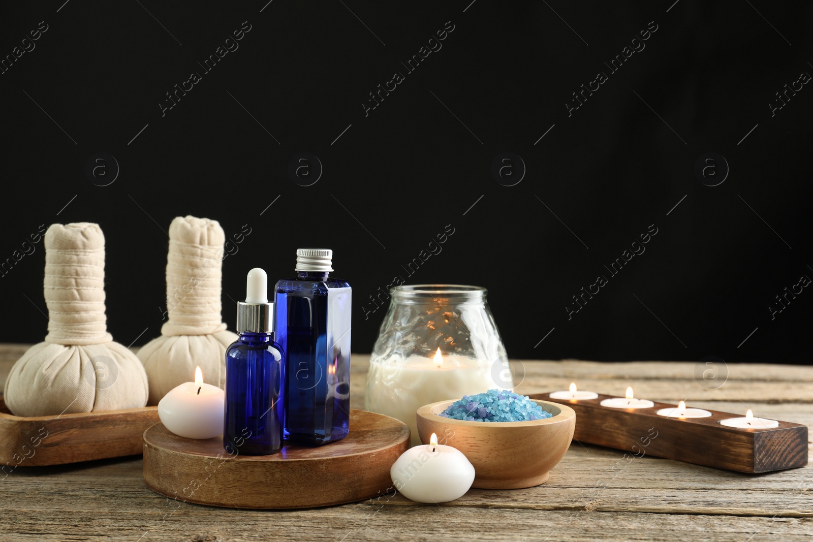 Photo of Spa composition. Cosmetic products, herbal bags, burning candles and sea salt on wooden table against black background, space for text