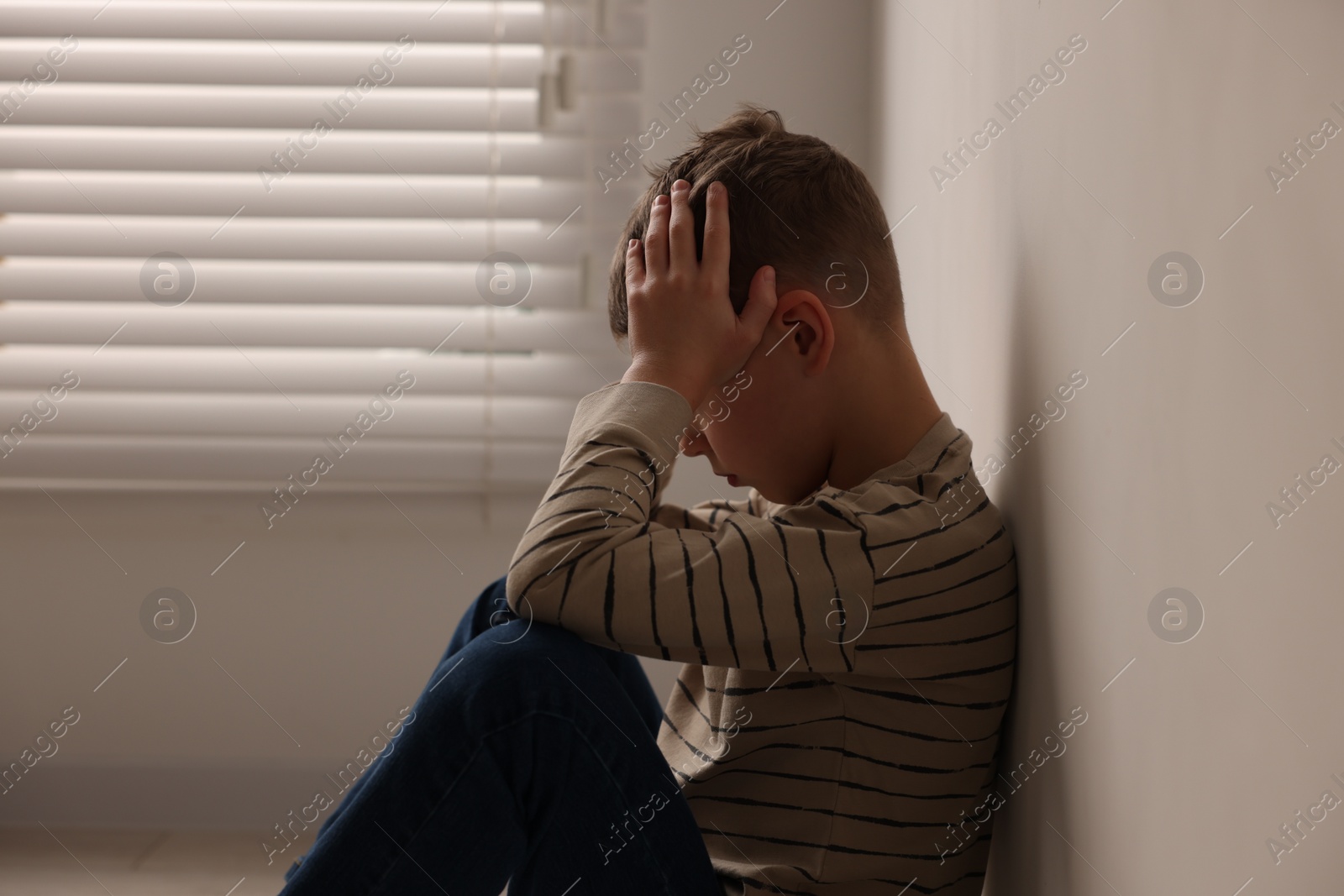 Photo of Child abuse. Upset boy sitting on floor near beige wall indoors