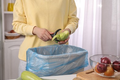 Photo of Woman peeling fresh zucchini above garbage bin indoors, closeup