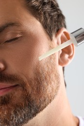 Photo of Handsome man applying cosmetic serum onto his face on light grey background, closeup