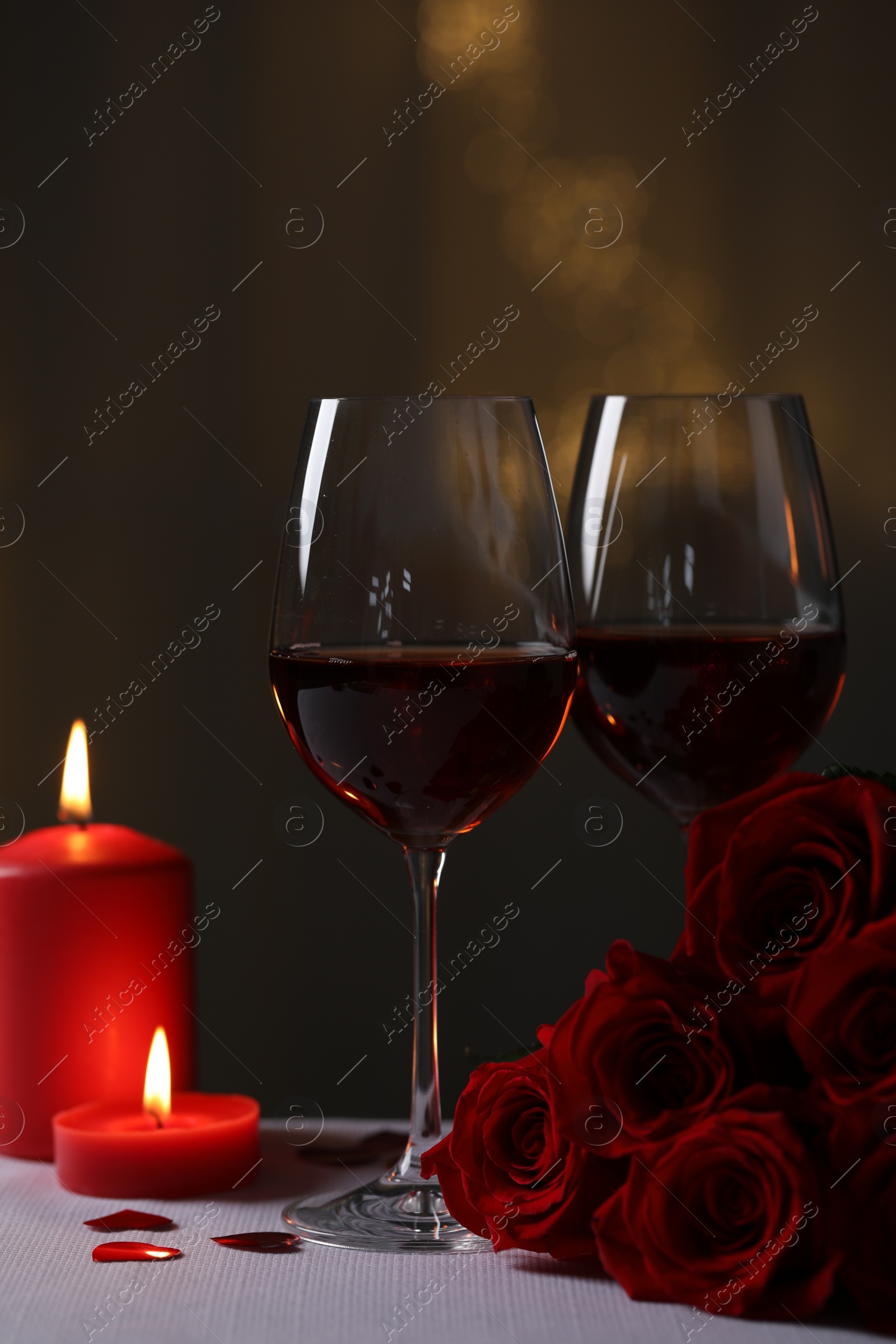 Photo of Glasses of red wine, burning candles and rose flowers on grey table against blurred lights. Romantic atmosphere