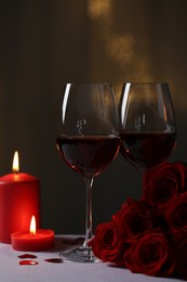 Glasses of red wine, burning candles and rose flowers on grey table against blurred lights. Romantic atmosphere