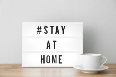Photo of Cup and lightbox with hashtag STAY AT HOME on wooden table near white wall. Message to promote self-isolation during COVID‑19 pandemic