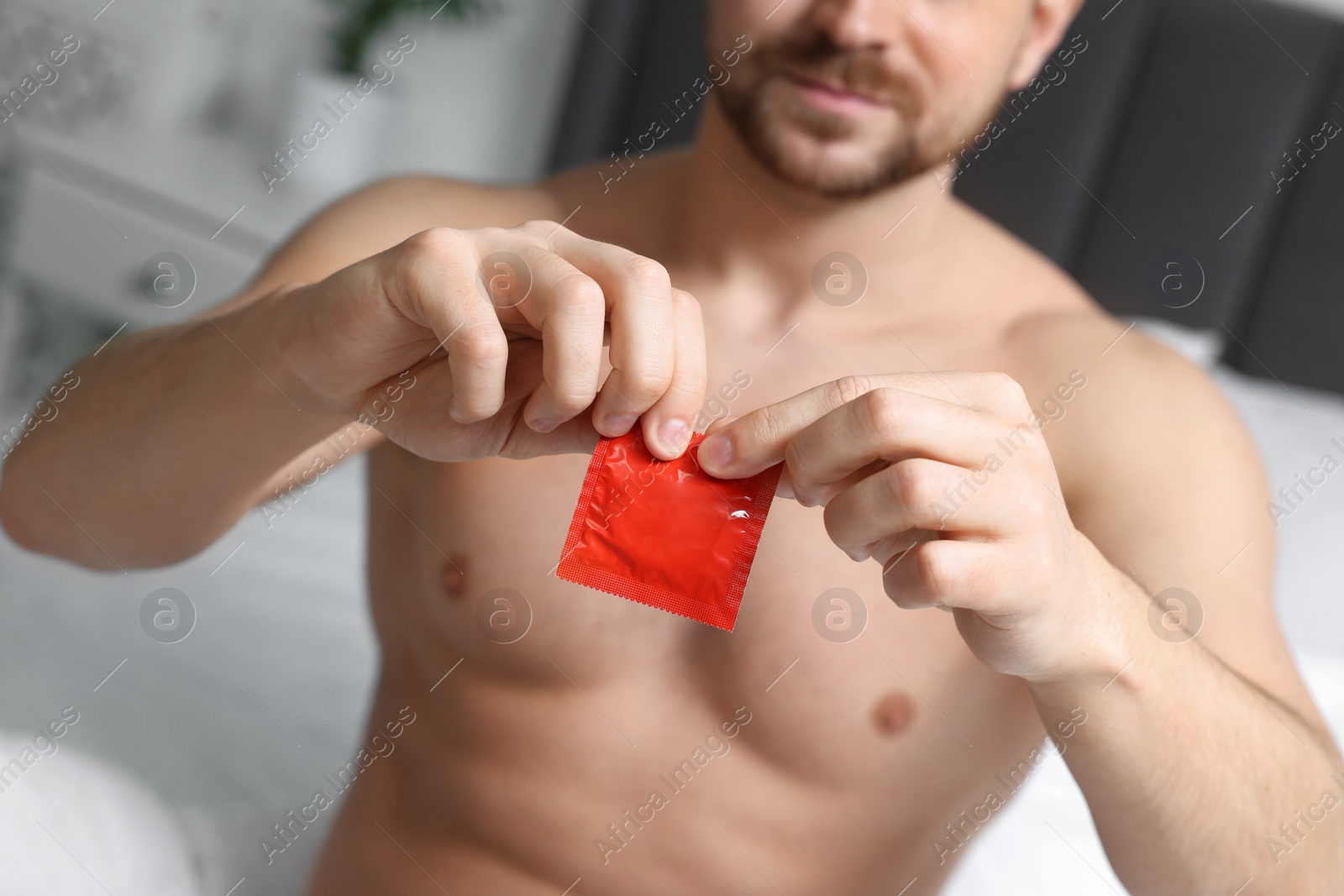 Photo of Closeup view of man opening pack with condom on bed indoors