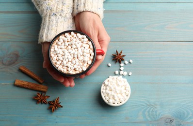 Woman with cup of tasty hot chocolate and marshmallows at light blue wooden table, top view. Space for text