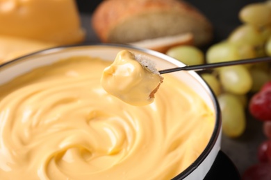 Photo of Piece of bread over pot with delicious cheese fondue, closeup