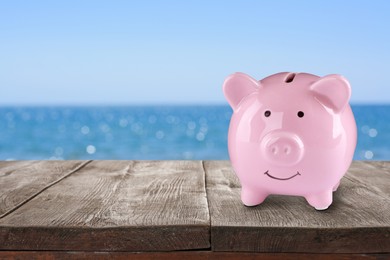 Image of Saving money for summer vacation. Piggy bank on wooden surface near sea, space for text