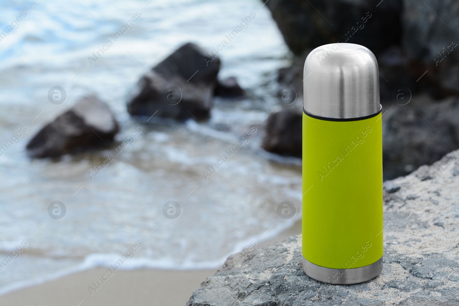 Photo of Metallic thermos with hot drink on stone near sea, space for text