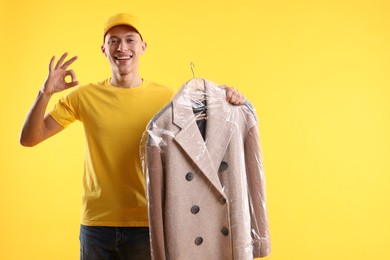 Photo of Dry-cleaning delivery. Happy courier holding coat in plastic bag and showing OK gesture on orange background, space for text