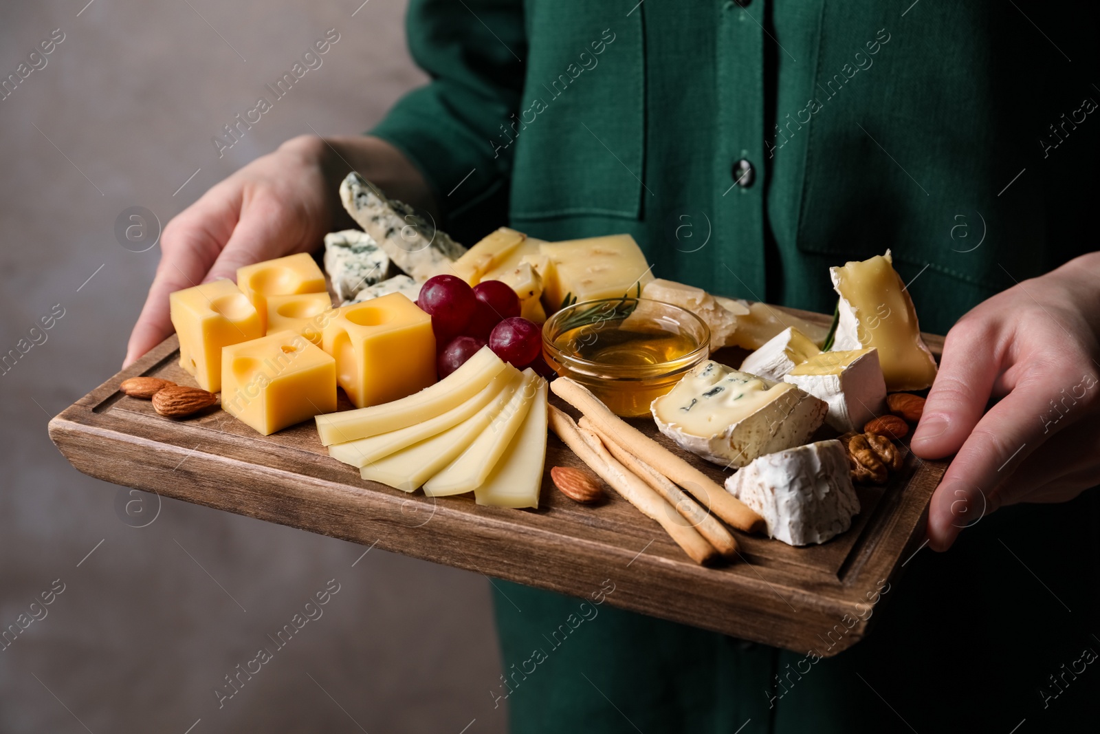 Photo of Woman holding cheese plate with honey, grissini and grapes on grey background, closeup