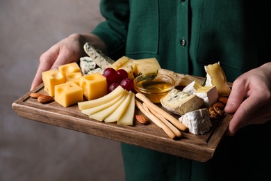 Photo of Woman holding cheese plate with honey, grissini and grapes on grey background, closeup