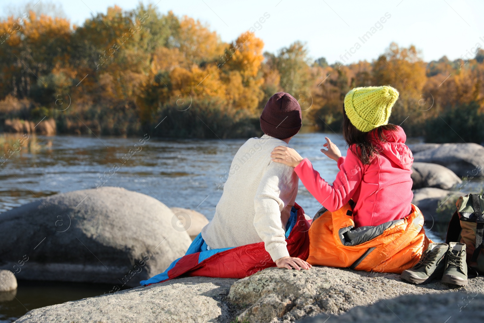 Photo of Couple of campers in sleeping bags sitting on rock near pond. Space for text