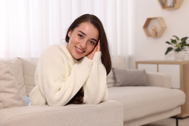 Photo of Beautiful young woman in stylish warm sweater on sofa at home, space for text