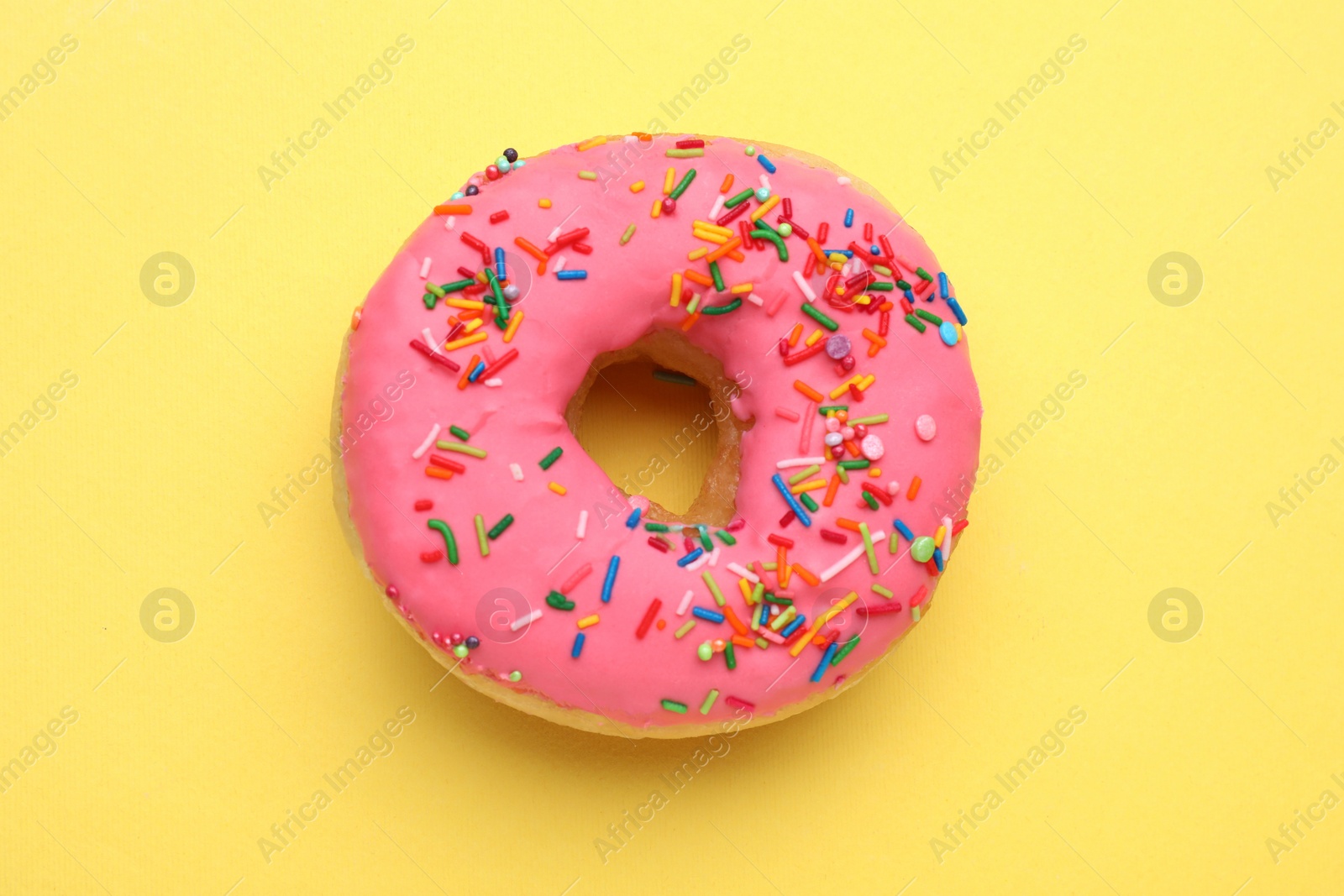 Photo of Glazed donut decorated with sprinkles on yellow background, top view. Tasty confectionery