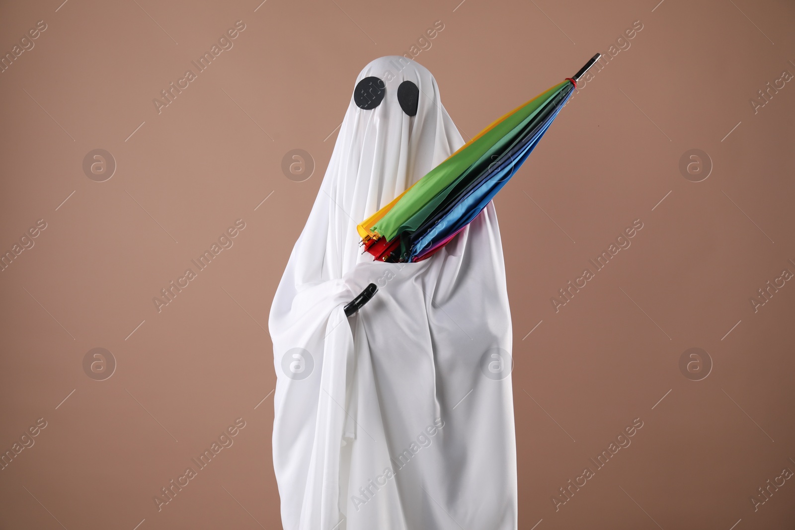 Photo of Person in ghost costume with rainbow umbrella on dark beige background