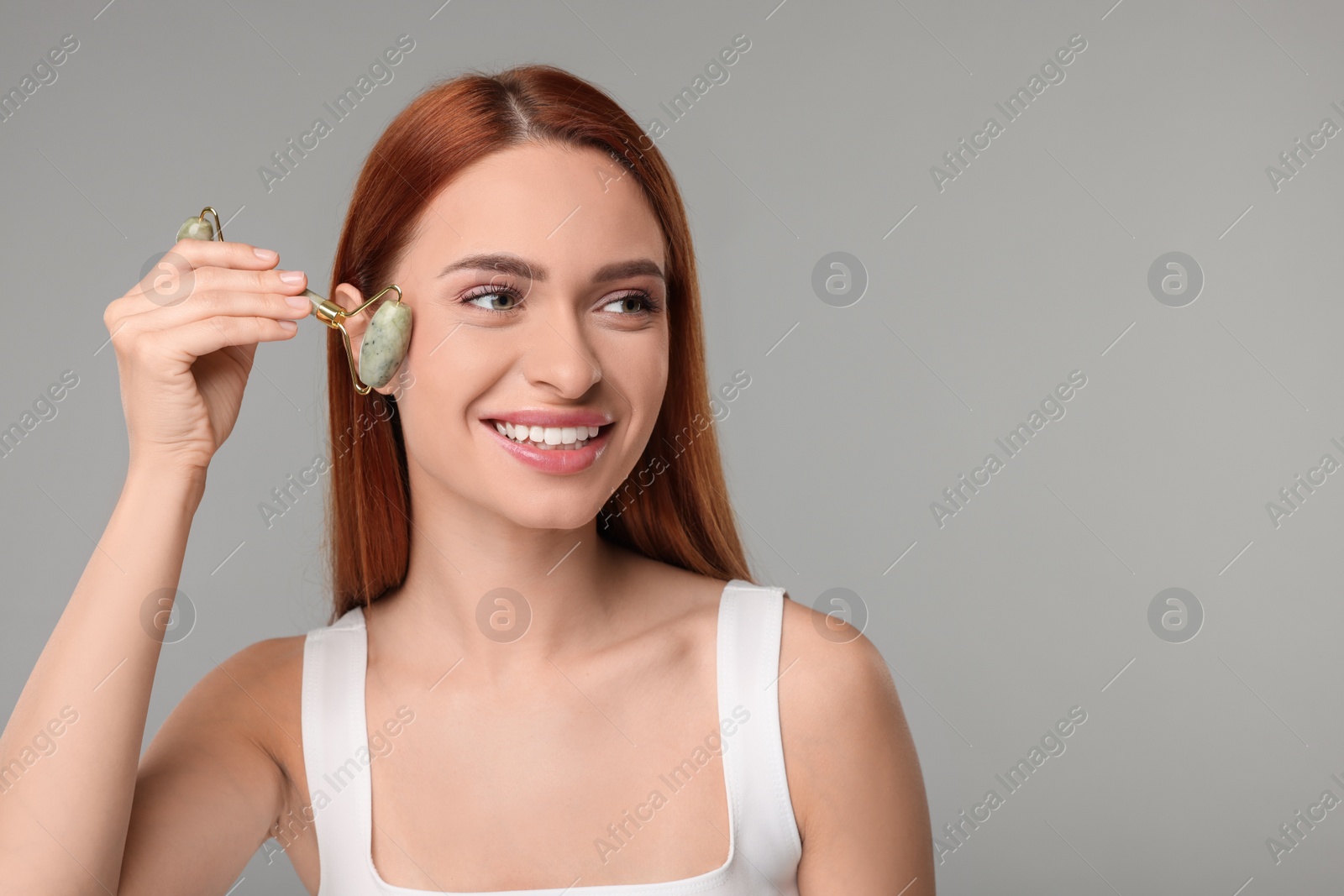 Photo of Young woman massaging her face with jade roller on grey background, space for text