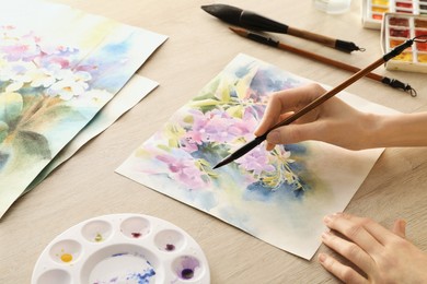 Photo of Woman painting flowers with watercolor at white wooden table, closeup. Creative artwork
