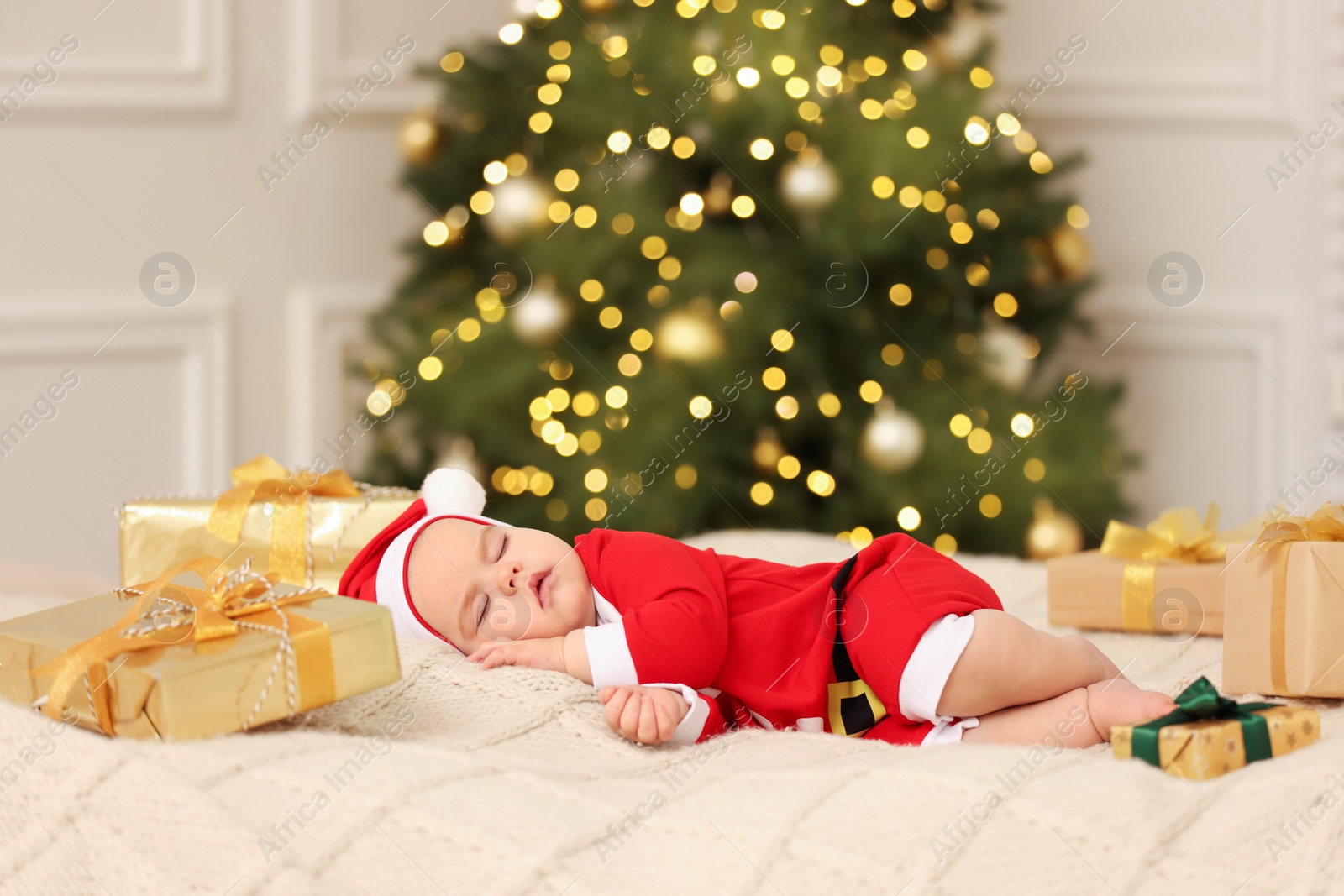 Photo of Cute baby in Christmas costume sleeping on knitted blanket against blurred festive lights. Winter holiday