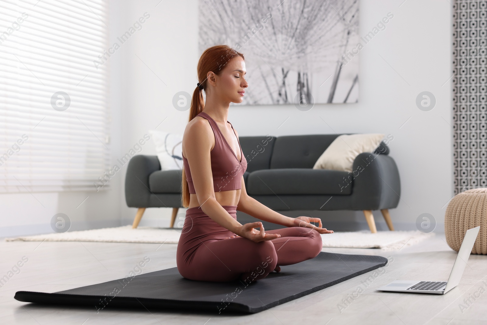 Photo of Beautiful young woman practicing Padmasana with laptop on yoga mat at home. Lotus pose